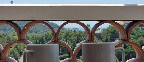 Two white ceramic cups on a wooden table with a metal railing in the foreground, overlooking a green landscape with trees and a clear sky.