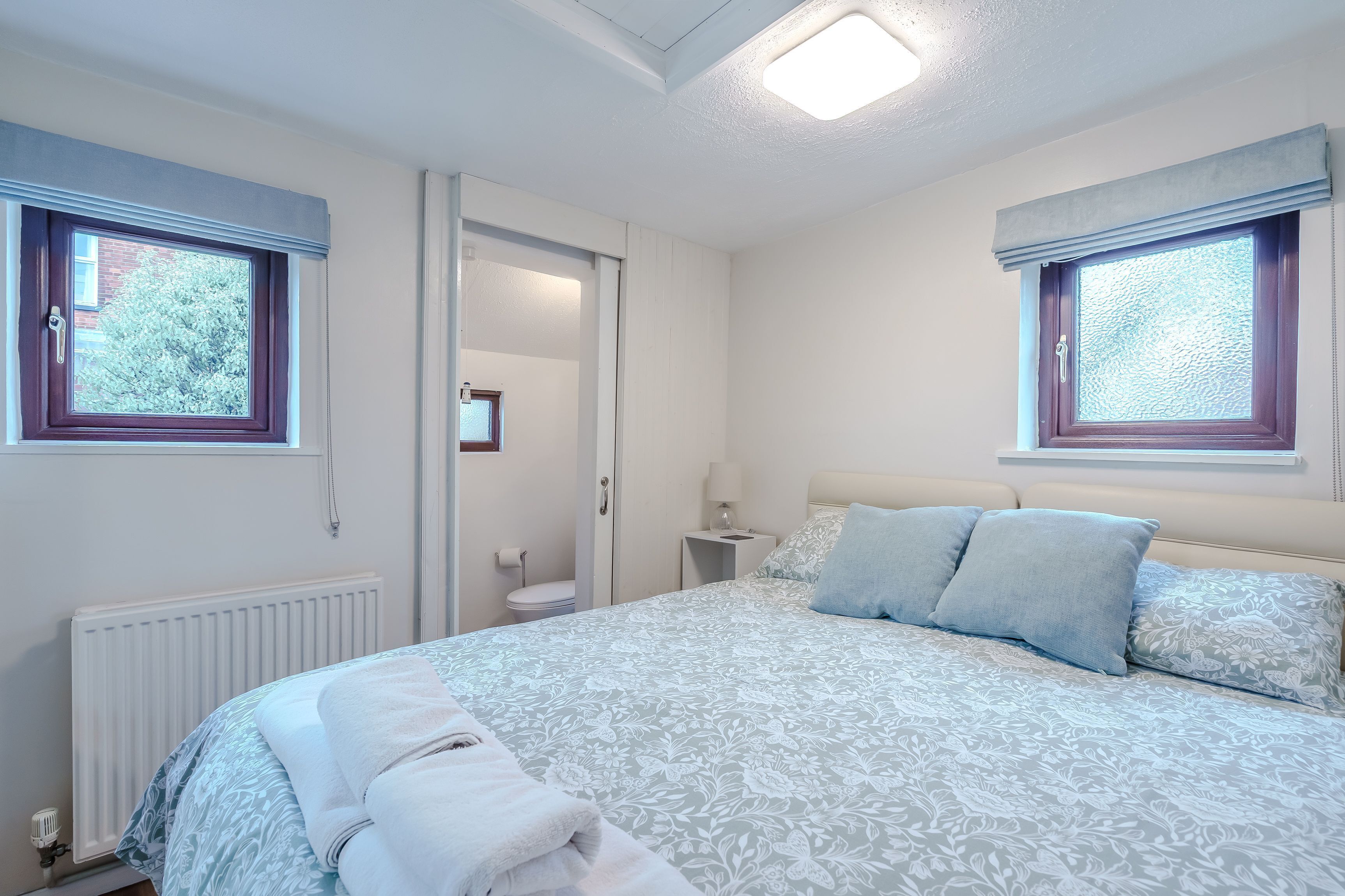 Un dormitorio con una cama, dos ventanas con cortinas, un radiador y una mesita de noche con lámpara.