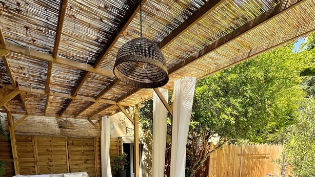 An outdoor patio with wooden furniture, a woven ceiling, and a view of the garden. - La Maison Près de la Fontaine (Talairan)