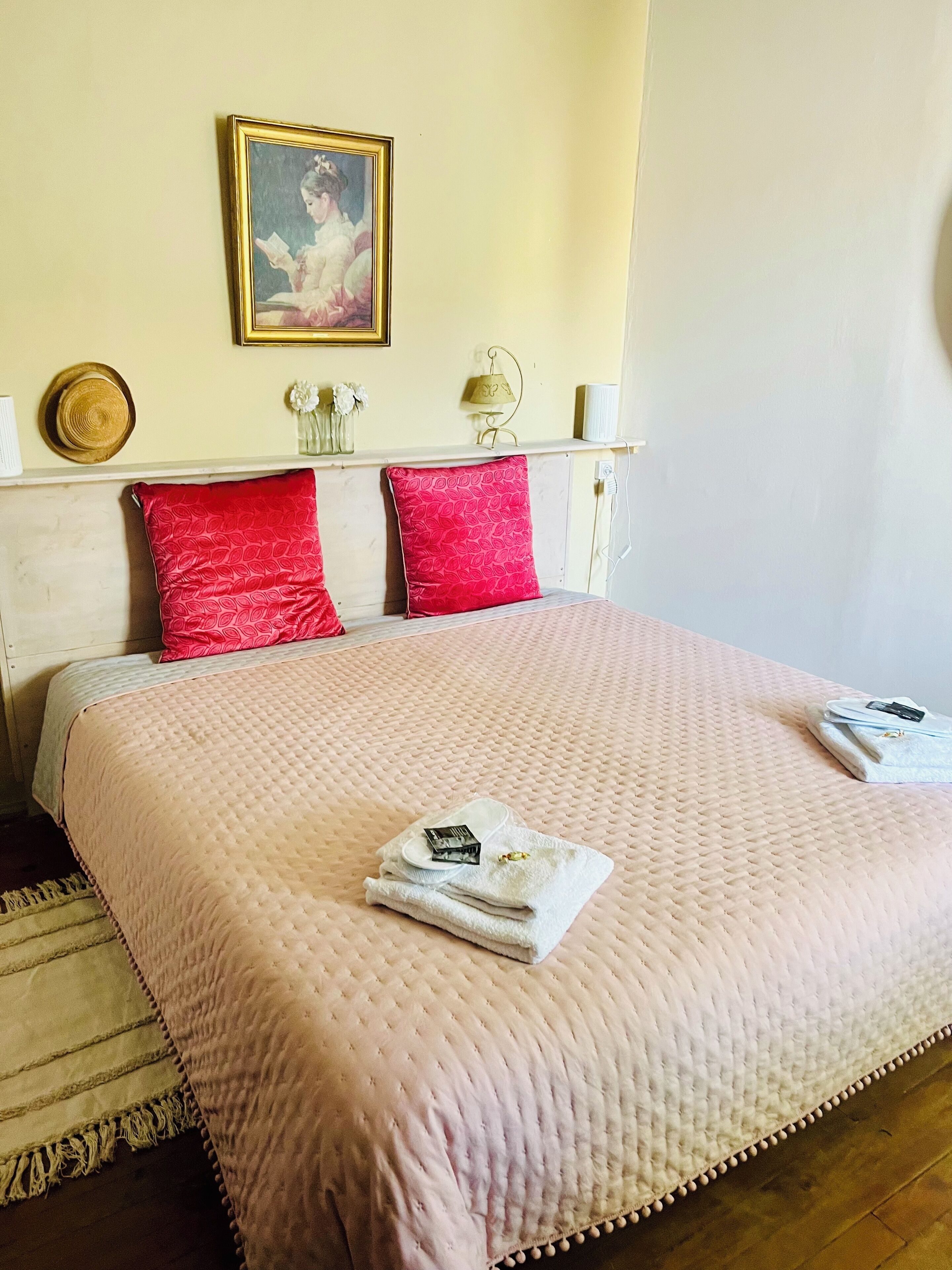 Un lit bien fait, recouvert d’une couverture rose, avec deux oreillers rouges et un tableau encadré accroché au mur.