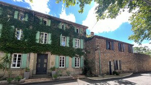 A two-story building with green shutters and ivy-covered walls, a stone building next to it, and a paved street in front. - La Maison Près de la Fontaine (Talairan)