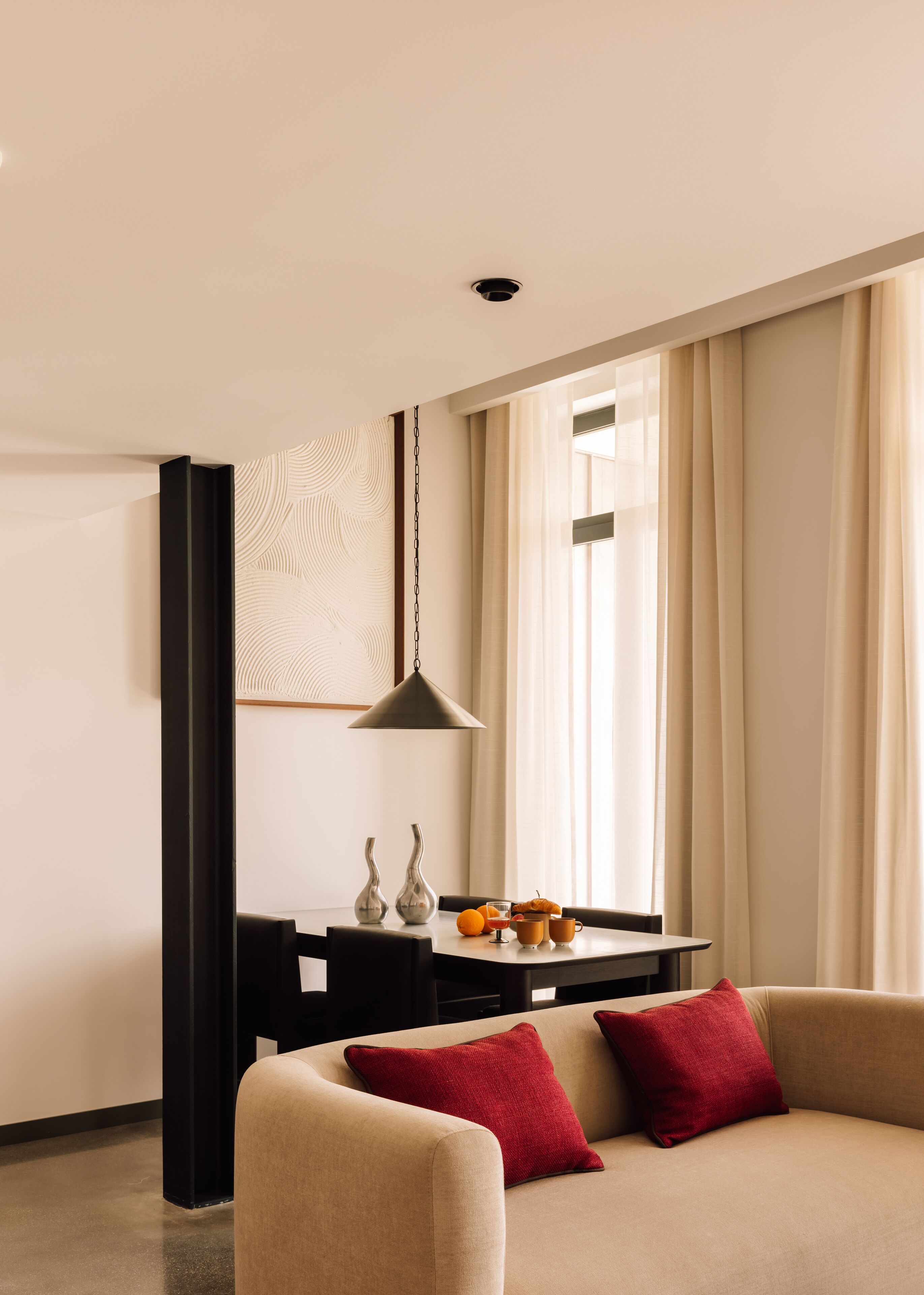 A modern living room with a beige sofa, red cushions, a dining table with fruit and glasses, and a pendant light.