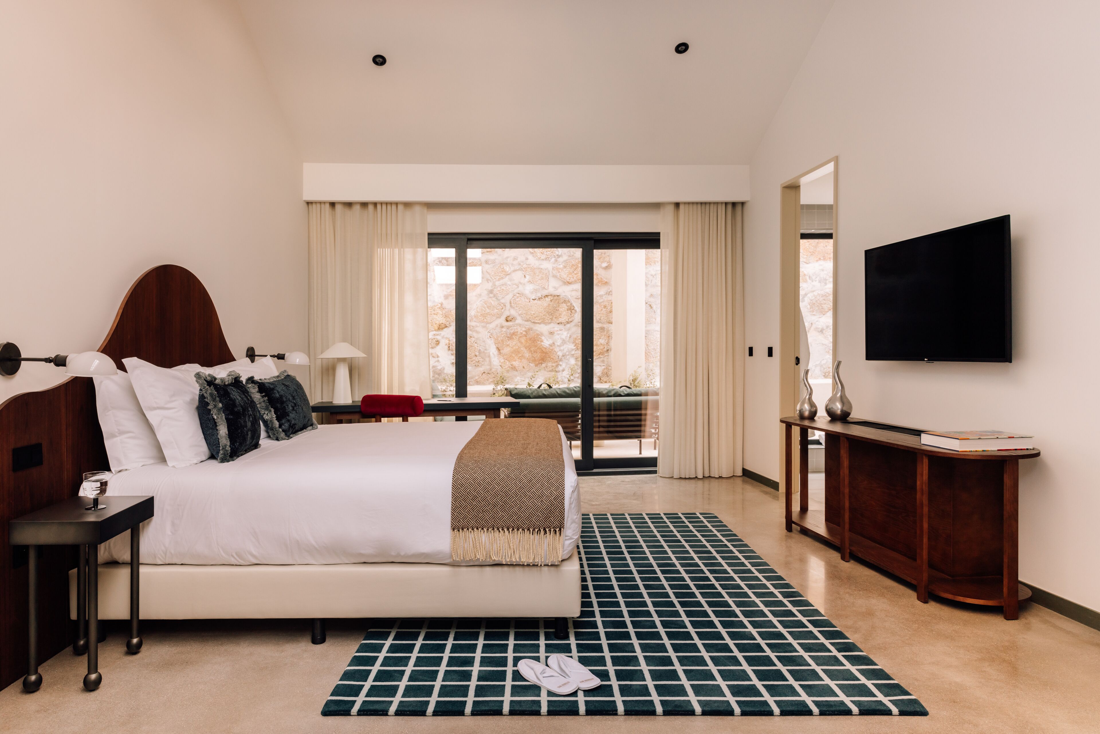 A modern hotel room with a large bed, a flat-screen TV, a small table, and a view of the outdoors through a large window.