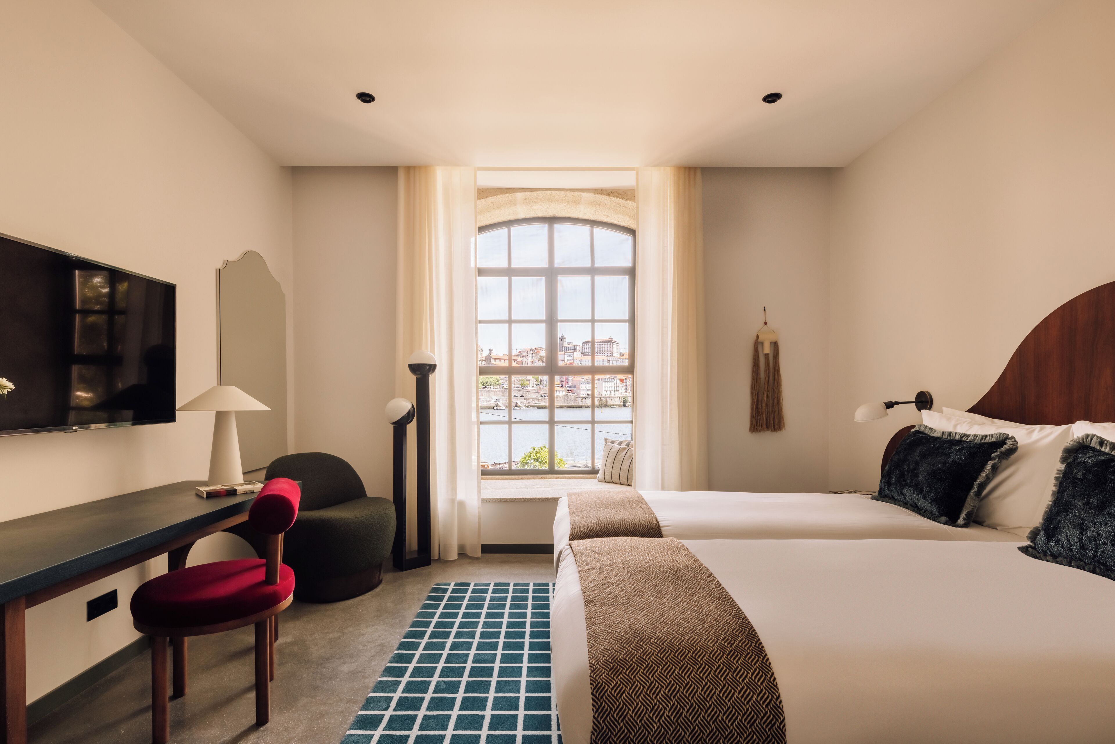 A hotel room with a bed, a desk, a chair, a TV, and a window with a view of buildings.