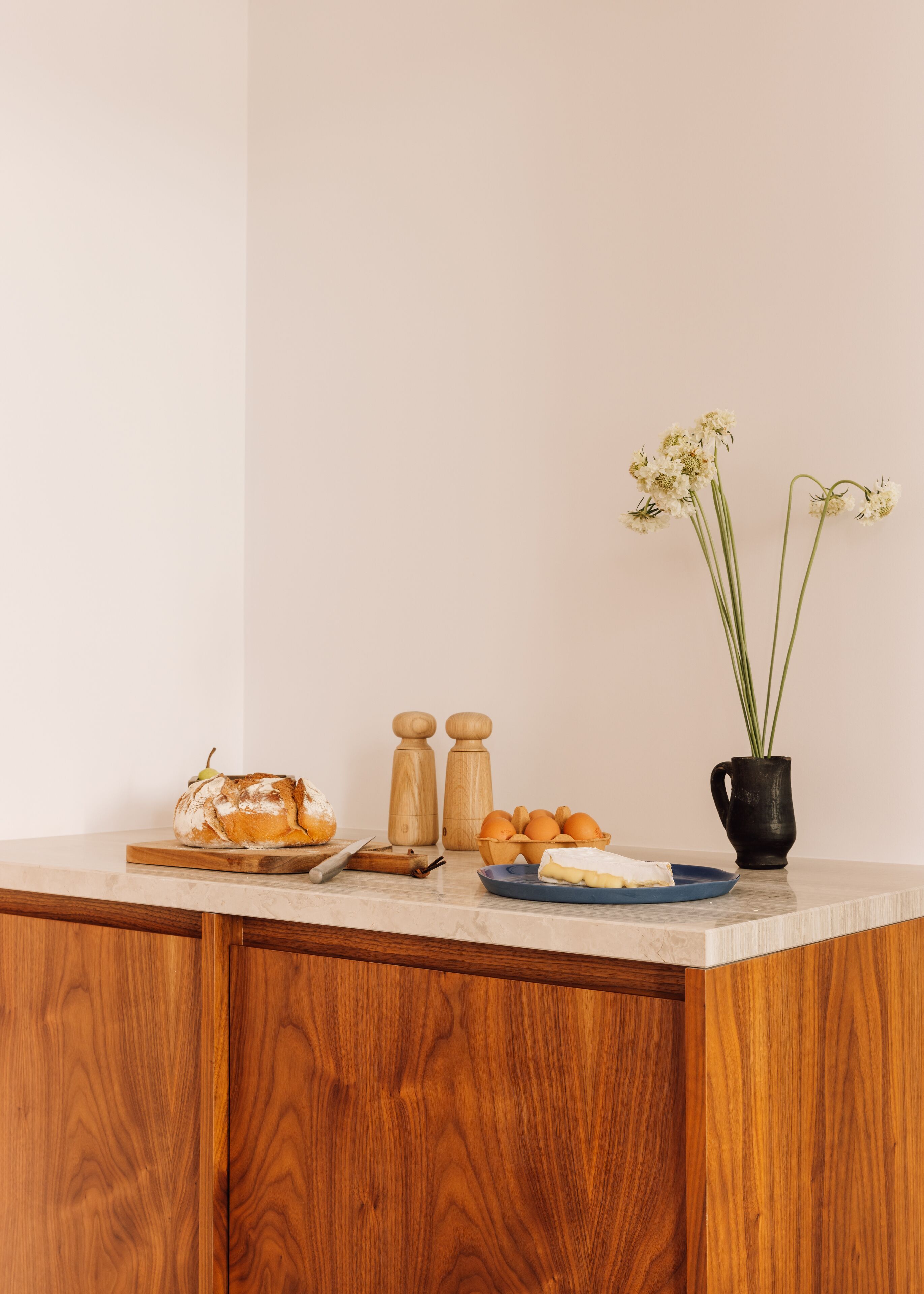 A kitchen counter with a loaf of bread, a bowl of oranges, a vase with flowers, and wooden salt and pepper shakers.
