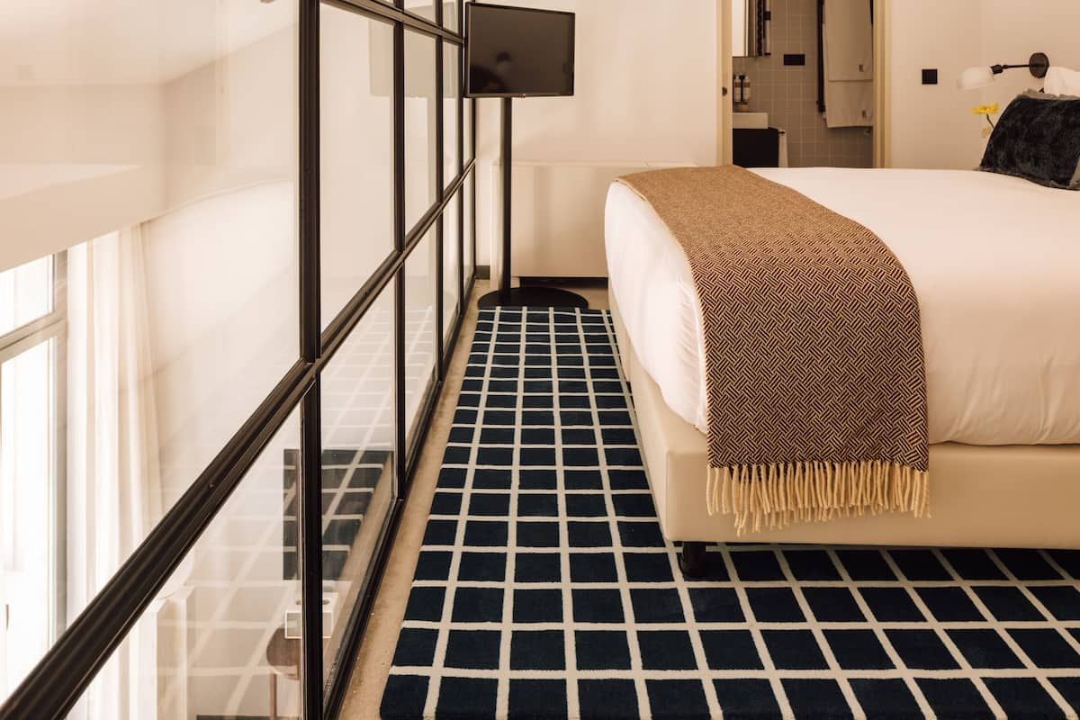 A modern hotel room with a large bed, a television, and a view through a glass wall.
