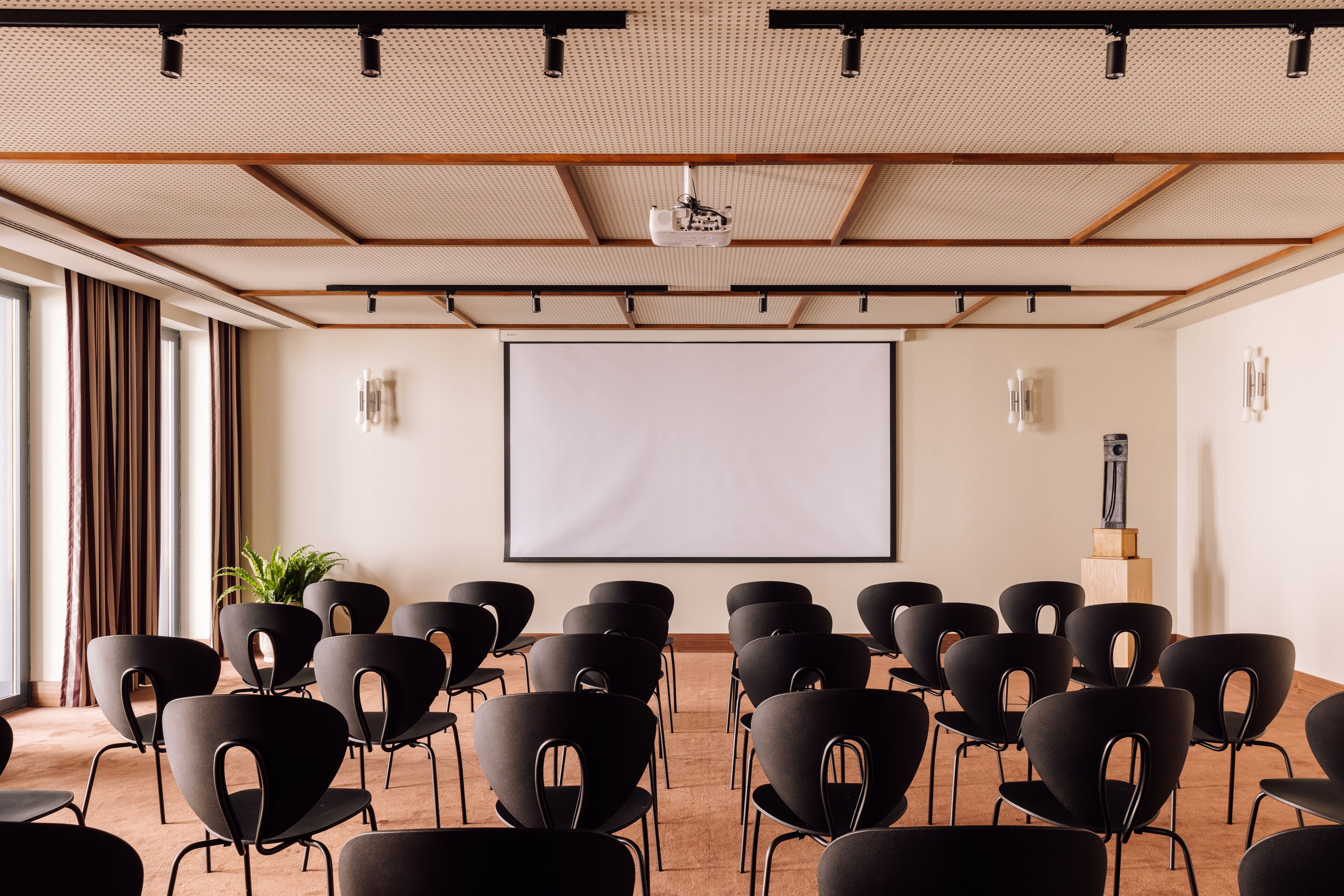 Un salón de conferencias con una pantalla grande, sillas negras, vigas de madera y una planta en maceta.