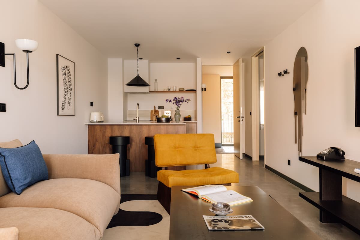 A modern living room with a sofa, a coffee table with a book, and a yellow chair. There is a kitchen area in the background with a bar stool and a hanging light fixture.