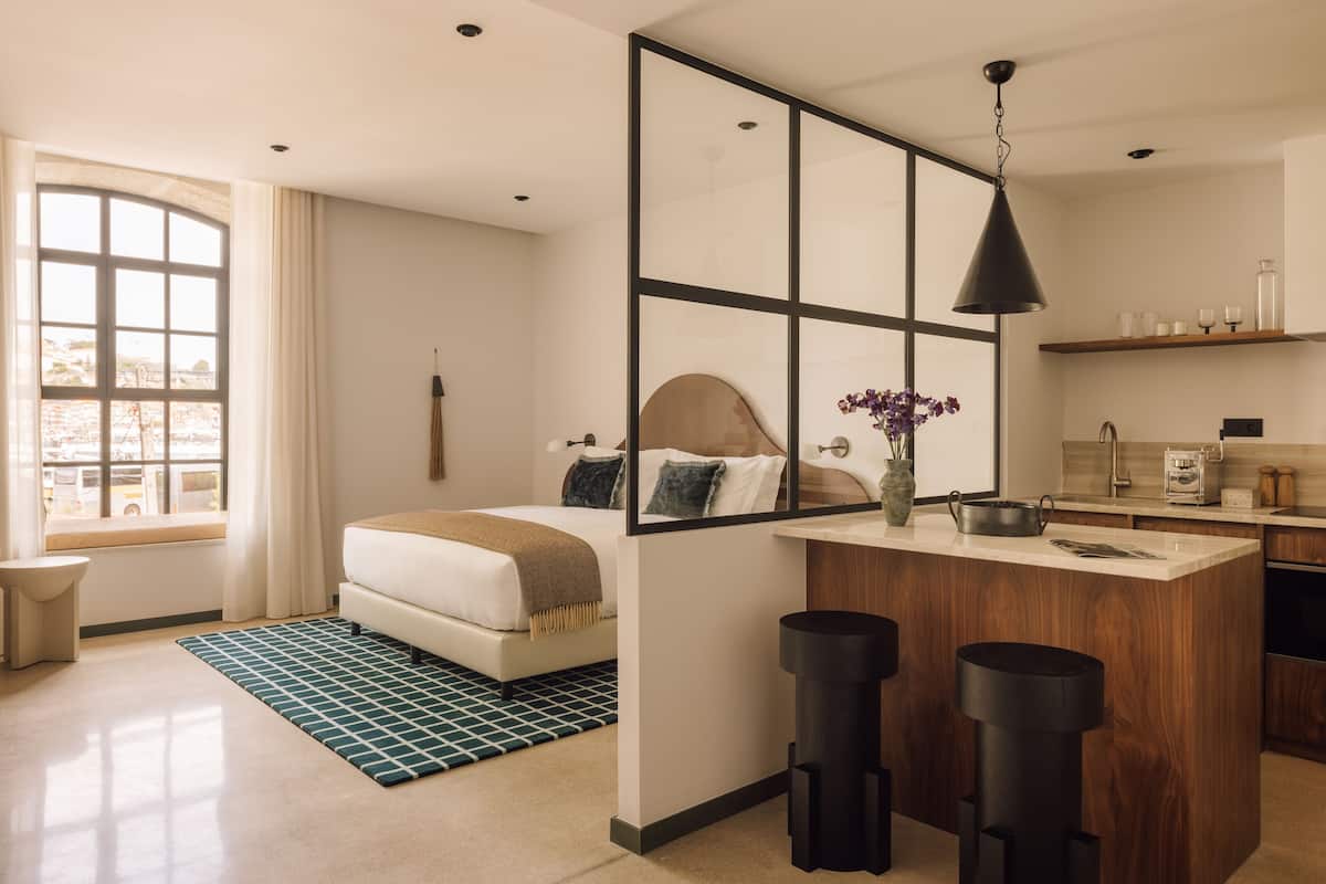A modern hotel room with a large bed, a small sitting area, a kitchenette, and a large window with curtains.