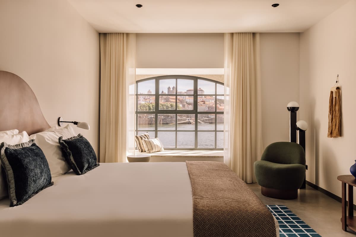 A hotel room with a large bed, a view of a river and buildings, a chair, and a lamp.