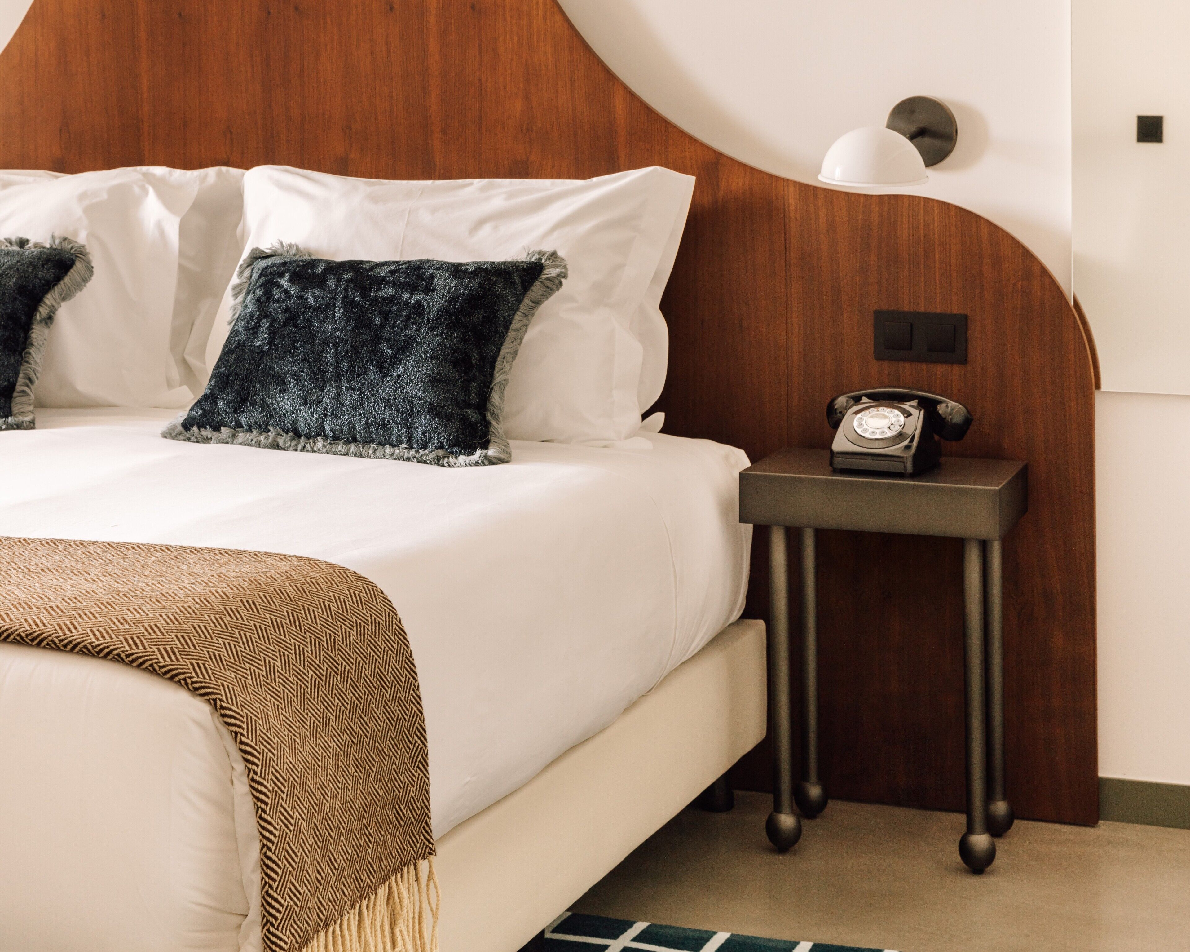 Una cama bien tendida con sábanas blancas y dos almohadas, una mesita de noche con teléfono giratorio y un cabecero de madera.