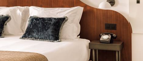Una cama bien tendida con sábanas blancas y dos almohadas, una mesita de noche con teléfono giratorio y un cabecero de madera.