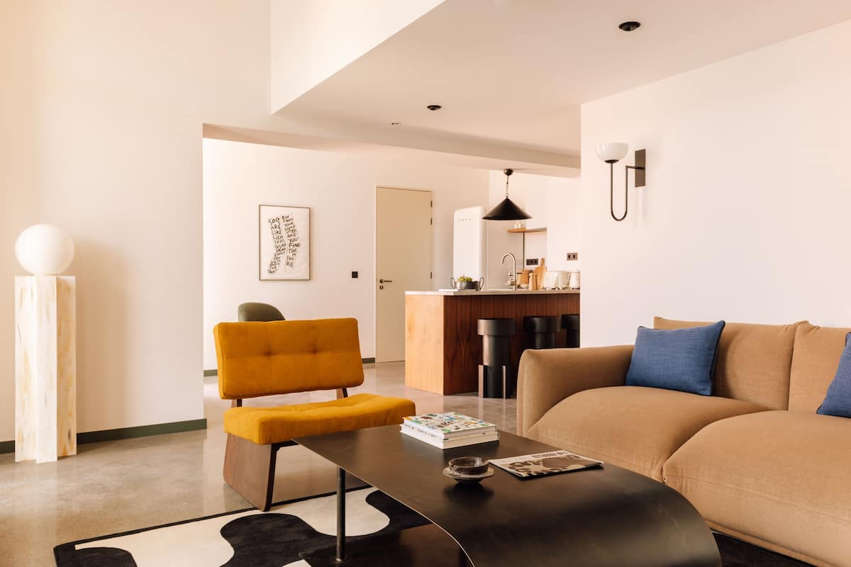 A modern living room with a sofa, a coffee table, and a yellow armchair. There is a kitchen area in the background with a refrigerator and a sink.