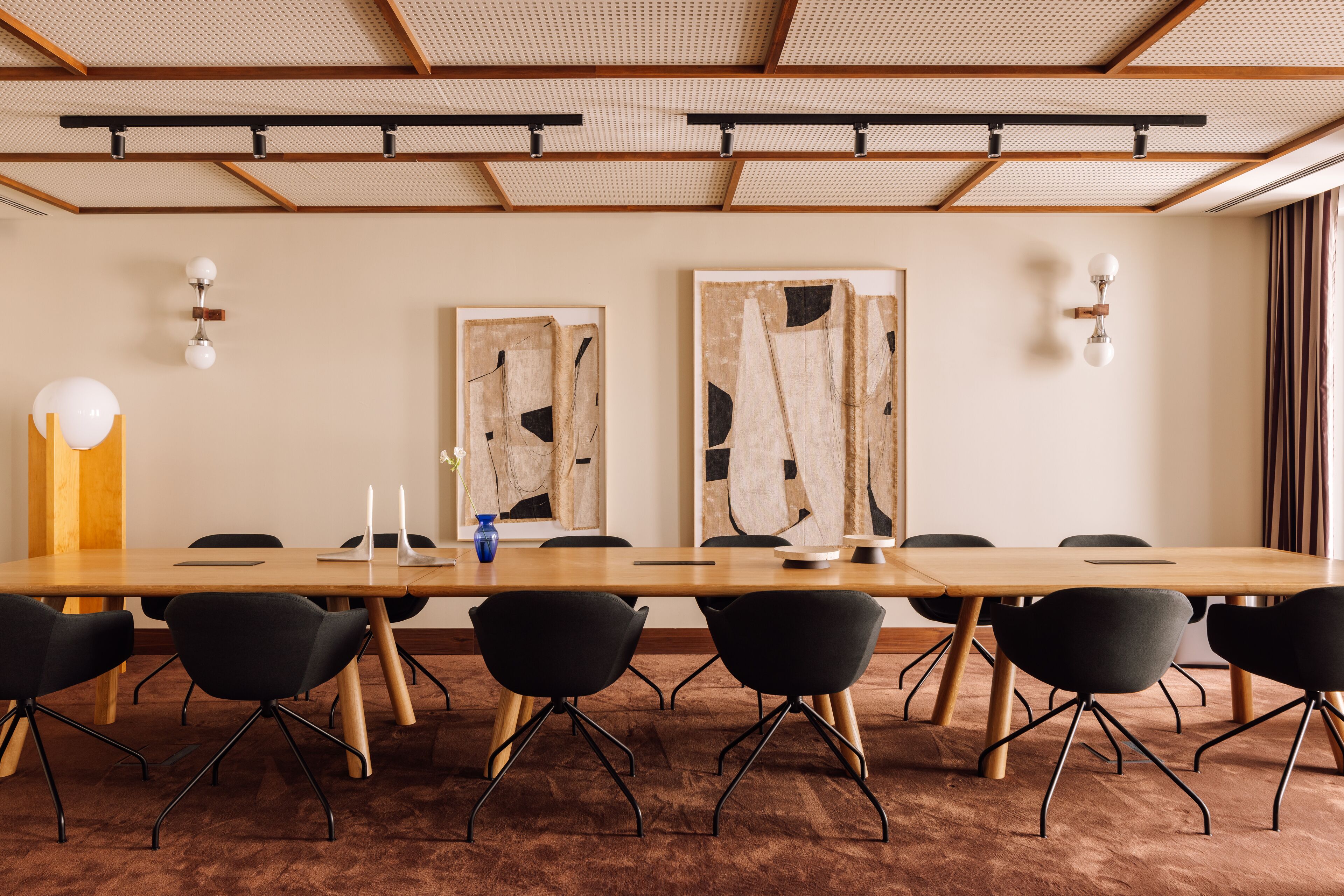 A conference room with a long wooden table, black chairs, abstract wall art, and a ceiling with wooden beams and track lighting.