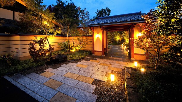 Een traditionele Japanse entree met een betegeld dak, houten pilaren en een pad van stenen dat leidt naar een lichtgevend pad.