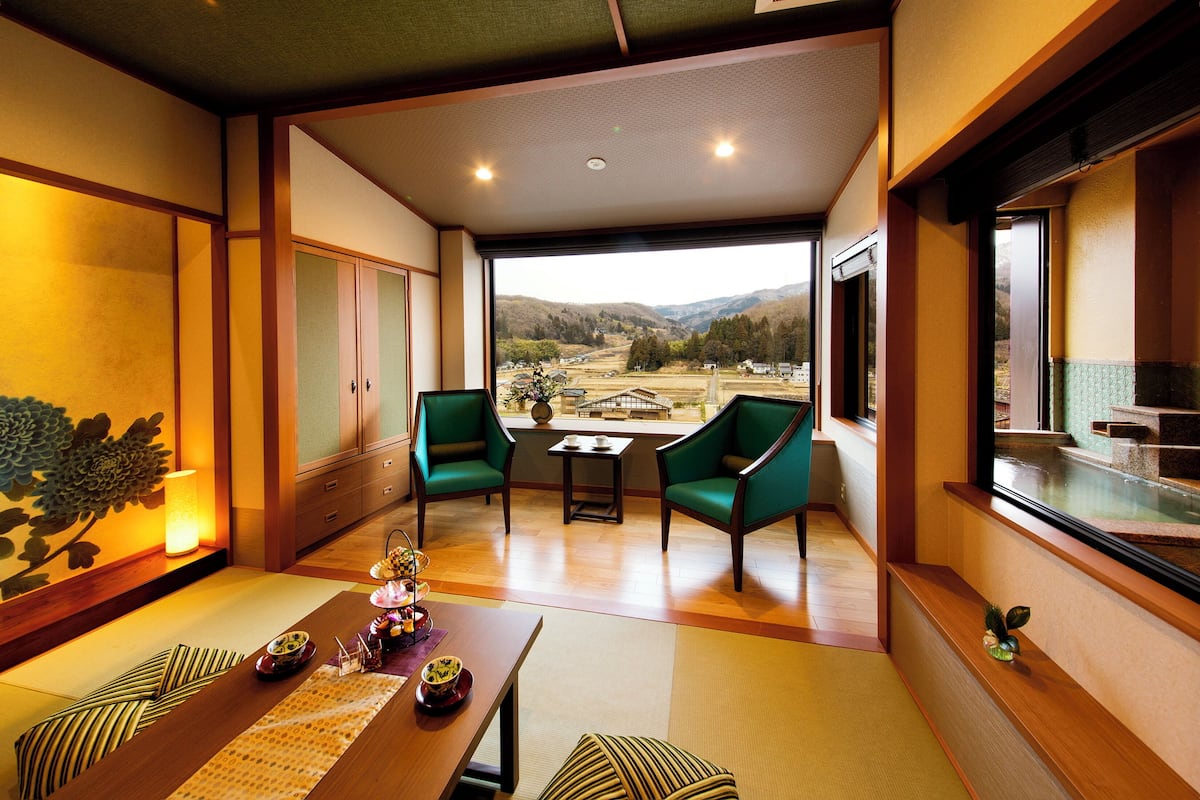 A traditional Japanese-style room with tatami flooring, a low table with a tea set, and a view of a mountainous landscape through large windows.