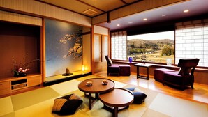 Een traditionele Japanse kamer met tatami vloer, een lage tafel met kussens, uitzicht op het platteland door grote ramen en een schuifdeur met een kersenbloesem schilderij.