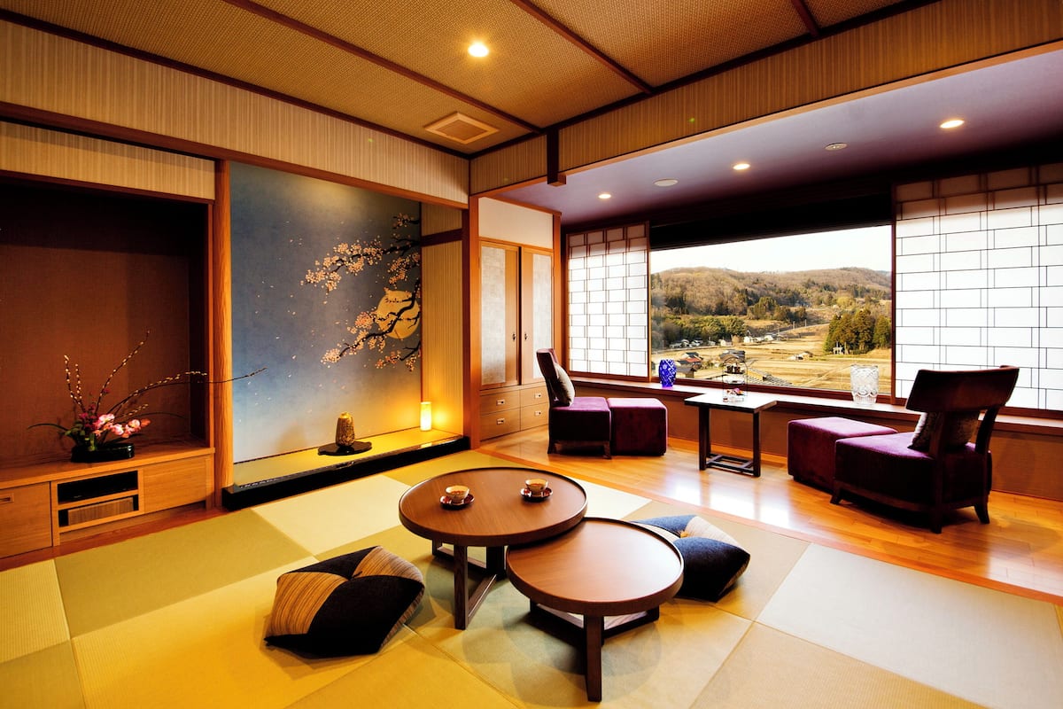 A traditional Japanese-style room with tatami flooring, a low table with cushions, a view of the countryside through large windows, and a sliding door with a cherry blossom painting.