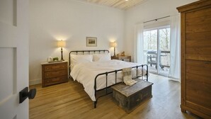 A bedroom with a wooden ceiling, a bed with white linens, two bedside tables with lamps, a wooden chest, and a view through a glass door to an outdoor area. - Pool, Pickleball & Basketball Court, Hot Tub, & Playset (Broken Bow)