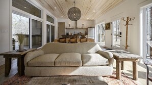 A modern living room with a large beige sofa, wooden ceiling, and large windows. - Pool, Pickleball & Basketball Court, Hot Tub, & Playset (Broken Bow)