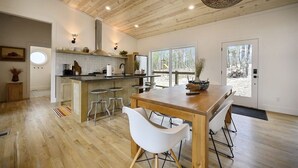 A modern kitchen with wooden beams, a central island with stools, and large windows offering a view of the outdoors. - Cabin w/ Heated Pool, Pickleball Court Access & Spacious Deck (Broken Bow)