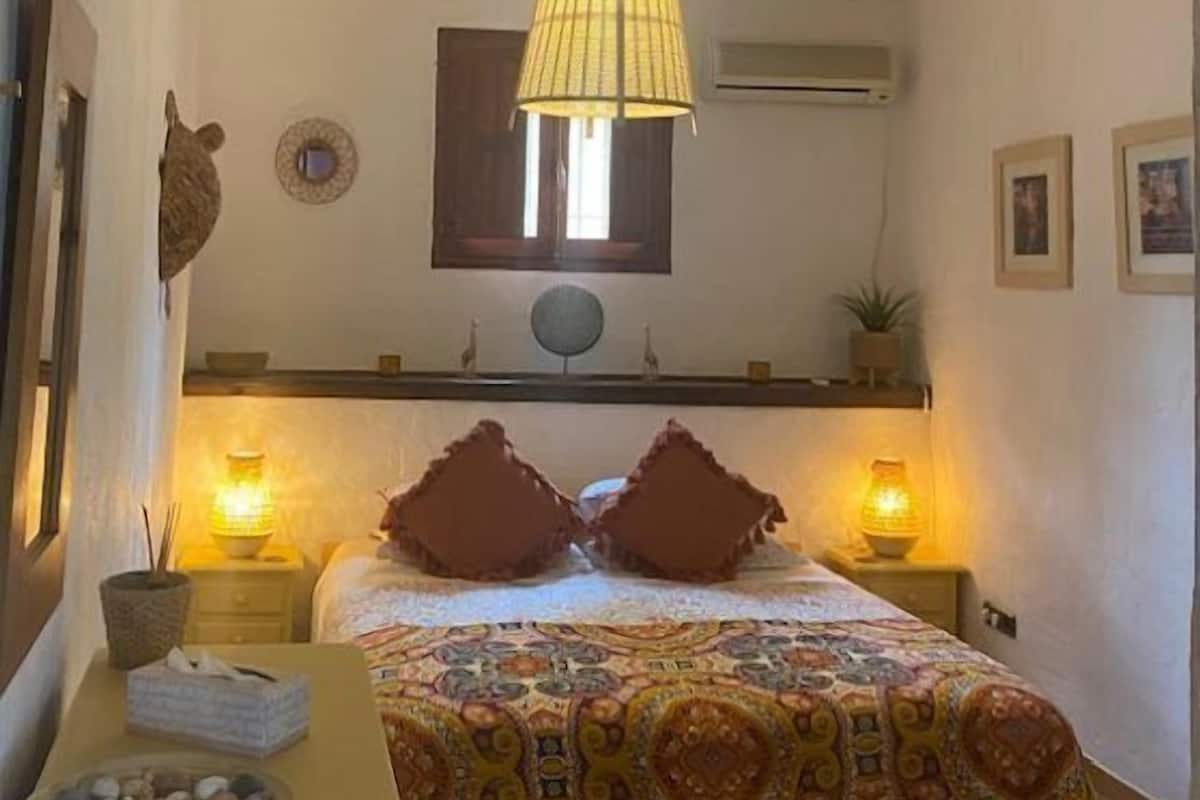 A bedroom with a bed, bedside lamps, a small table with a bowl of stones, and a hanging light fixture.