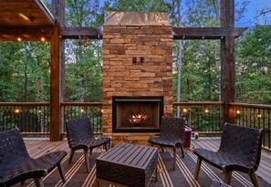 Une terrasse extérieure avec une cheminée en pierre, des meubles en bois et une vue sur les arbres.