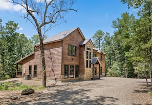 A large wooden cabin with a spacious porch and a gravel driveway, surrounded by trees.