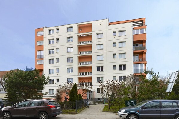 A multi-story residential building with balconies, a gated entrance, and a parking area with cars.