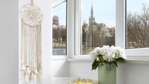 Una habitación de esquina con un ventanal grande, un escritorio blanco con un jarrón de flores, un tapiz macramé y una silla blanca.
