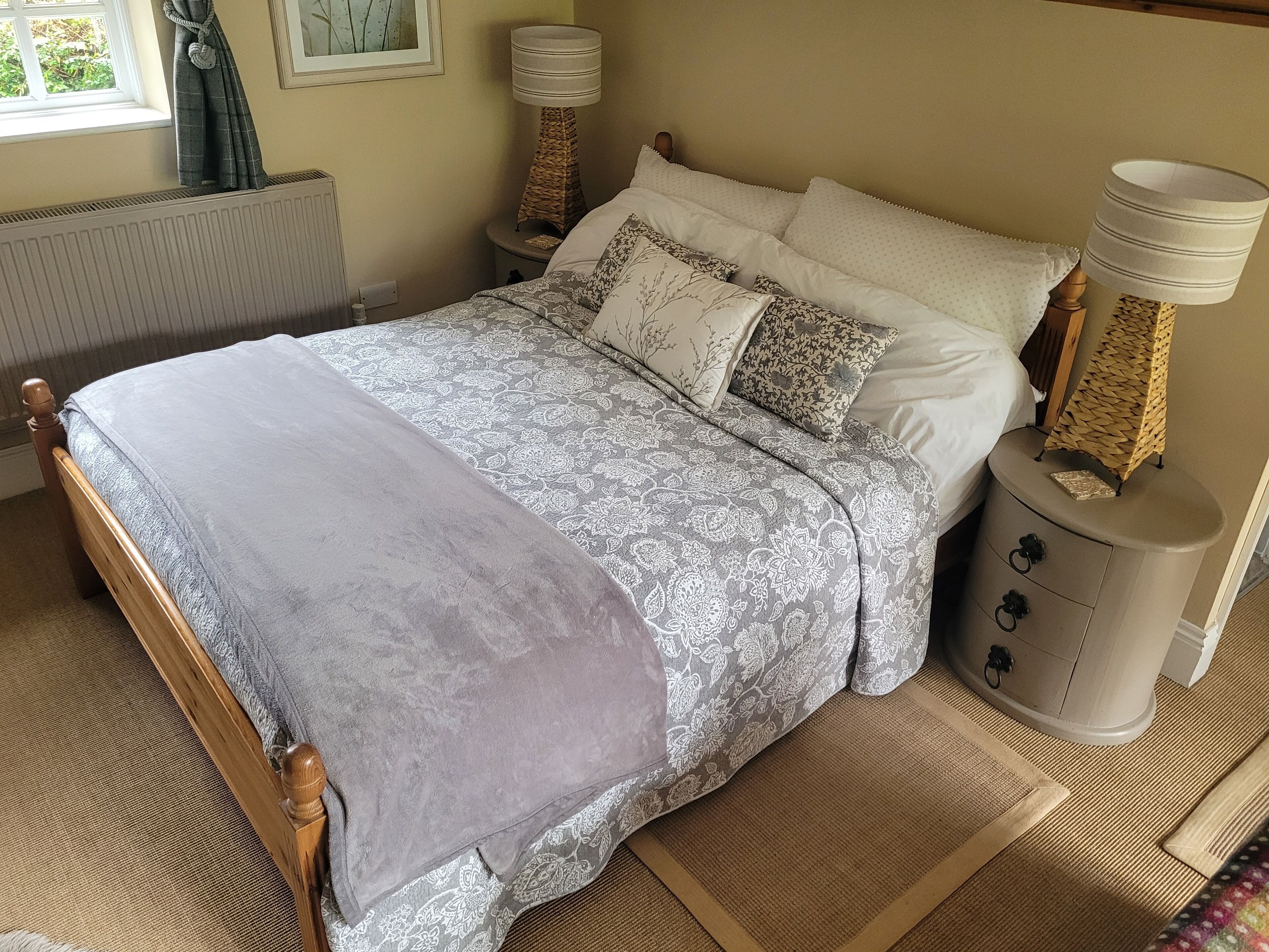 Ein ordentlich gemachtes Bett mit grauer Decke, ein Nachttisch mit Lampe und ein Fenster mit Vorhängen.