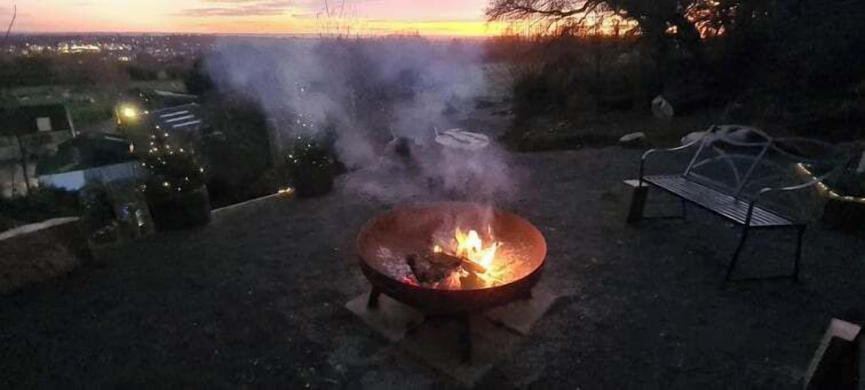A fire pit with flames, surrounded by a metal chair and a table, set on a concrete surface.