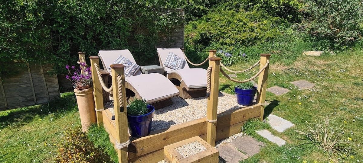 A wooden deck with three lounge chairs and a small table, surrounded by potted plants and a stone pathway.