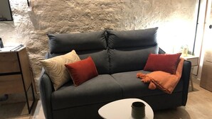 Un canapé gris foncé agrémenté de plusieurs coussins, une petite table blanche surmontée d’un récipient foncé et un plancher en bois.