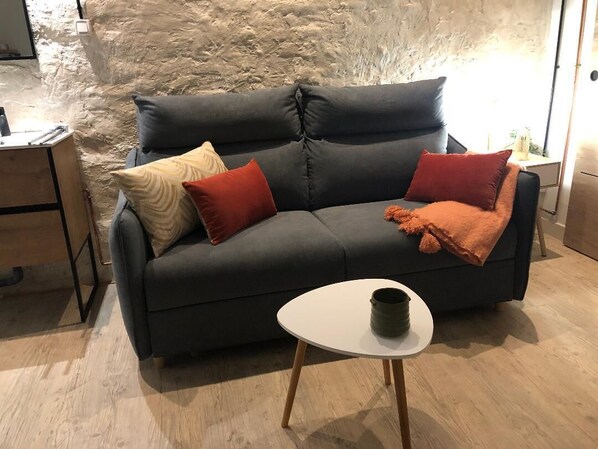 A dark grey sofa with multiple cushions, a small white table with a dark container, and a wooden floor.