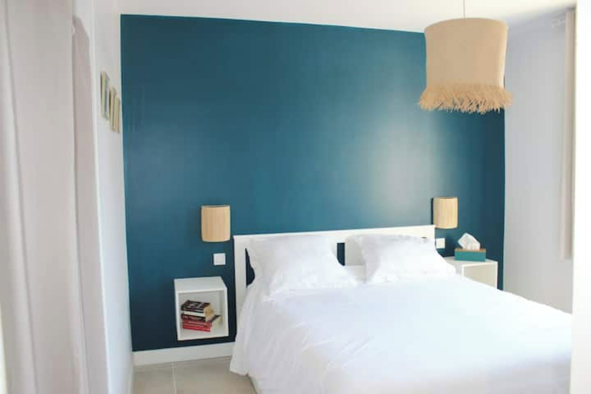 Ein Schlafzimmer mit einem weißen Bett, zwei Nachttischlampen, einem kleinen Nachttisch mit einem Buch und einer Wandlampe.