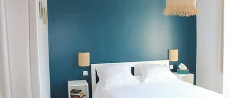 Ein Schlafzimmer mit einem weißen Bett, zwei Nachttischlampen, einem kleinen Nachttisch mit einem Buch und einer Wandlampe.