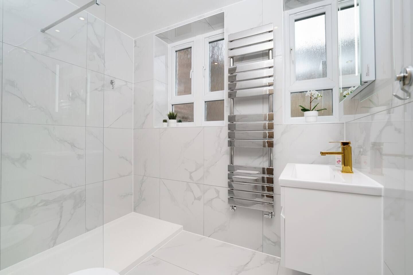 A modern bathroom with a glass shower enclosure, a white marble floor, a gold faucet, and a white sink.