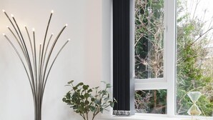 Quarto moderno com luminária de chão alta, uma mesa pequena com planta em vaso e janela com vista para árvores.