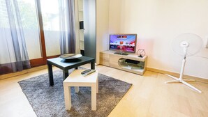 Un salon moderne avec un téléviseur à écran plat, un ventilateur sur pied, une petite table avec une télécommande et un tapis gris.