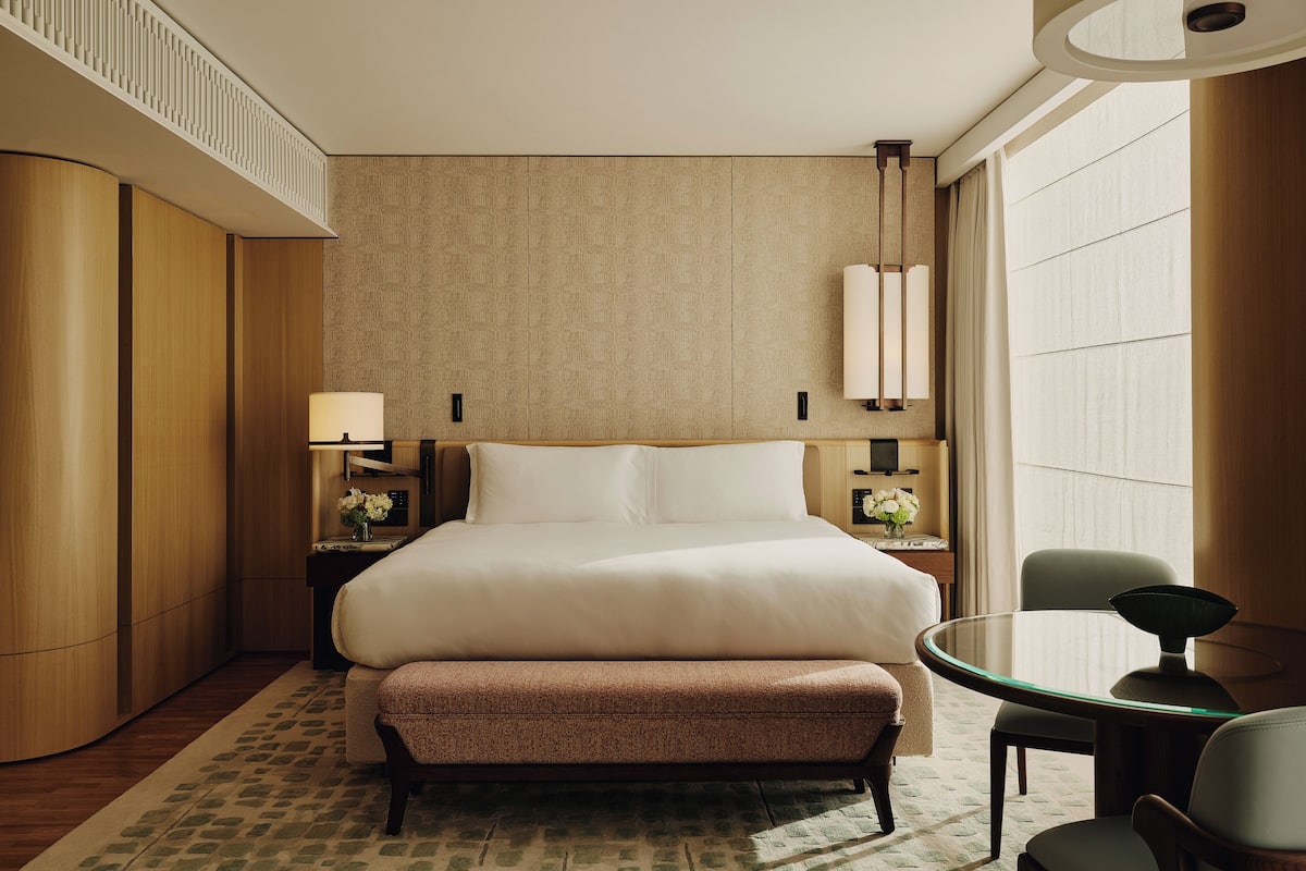 A hotel room with a large bed, a round glass table, a chair, a nightstand with a lamp, and a wall-mounted light fixture.