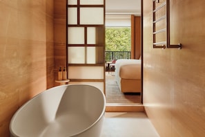 A modern bathroom with a freestanding bathtub, a small table with toiletries, and a view of a balcony with greenery. - The Emory, Maybourne (London)