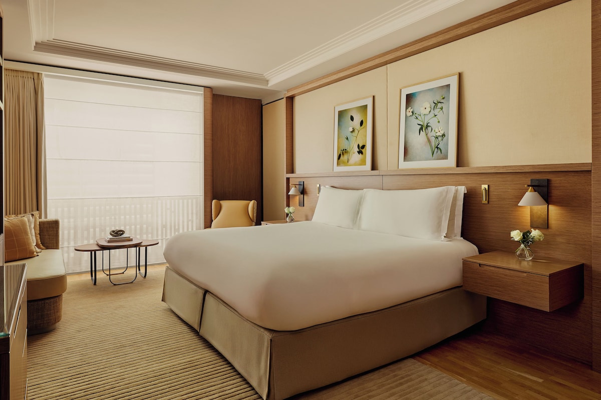 A hotel room with a large bed, a small table, a chair, and artwork on the wall.