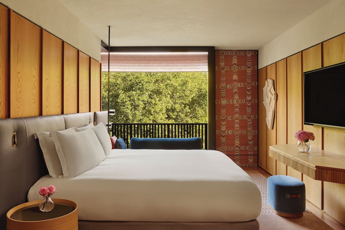 A modern hotel room with a large bed, a small desk, a TV, and a balcony with a view of trees.