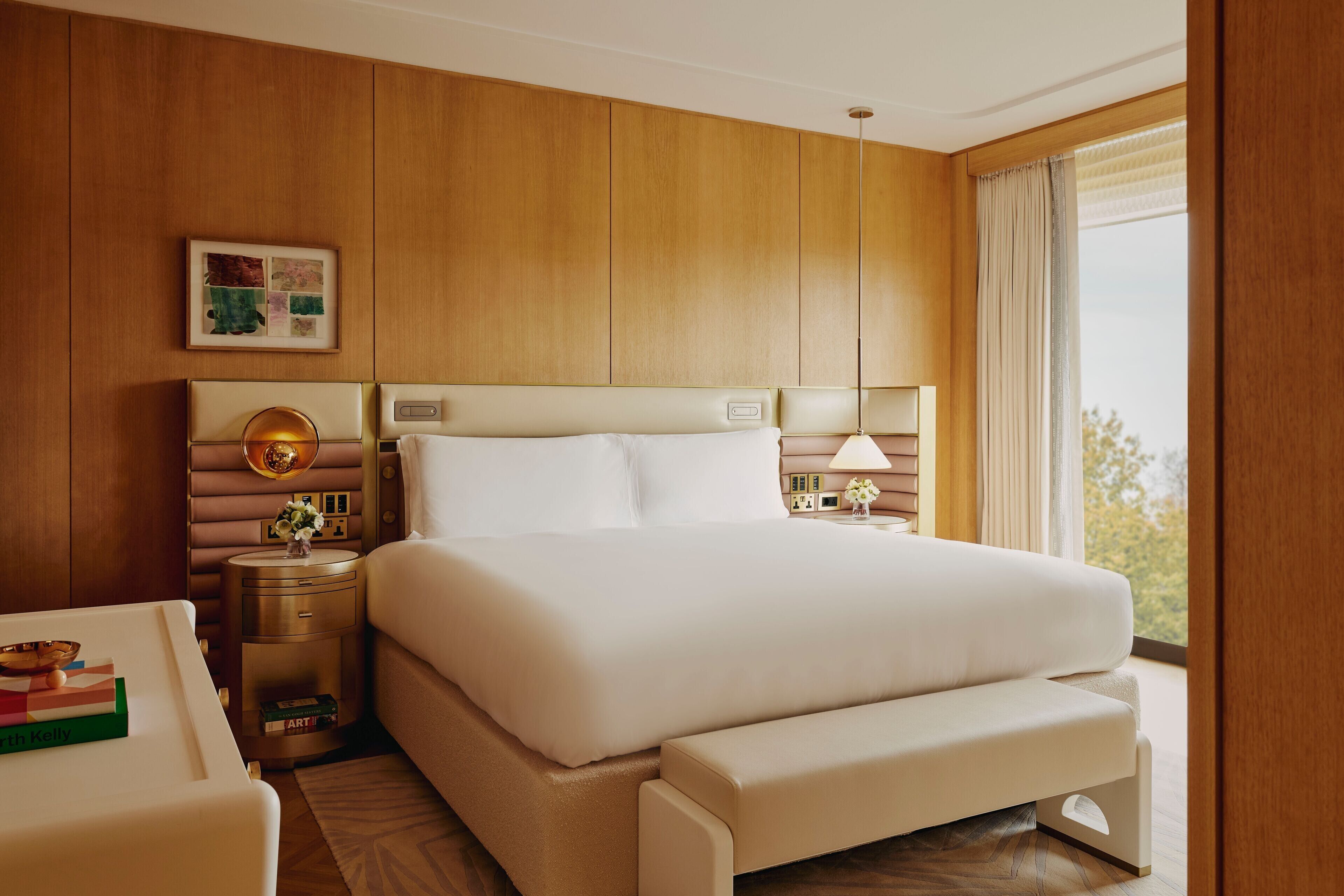 Une chambre d’hôtel avec un grand lit, des tables de chevet, une petite table, un banc, un tableau encadré et une fenêtre avec des rideaux.