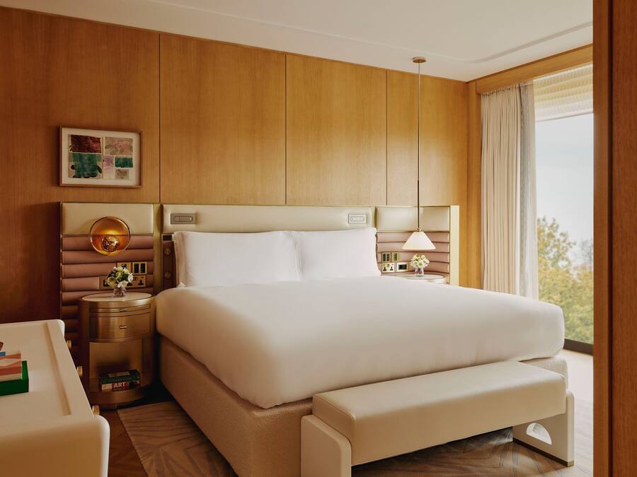 Una habitación de hotel con una cama grande, mesitas de noche, un banco, un cuadro enmarco y una ventana con cortinas.