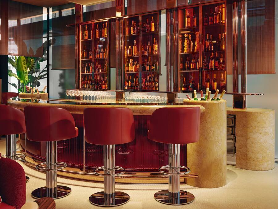 Een bar met een houten toog, rode barstoelen en gevulde flessenrekken.