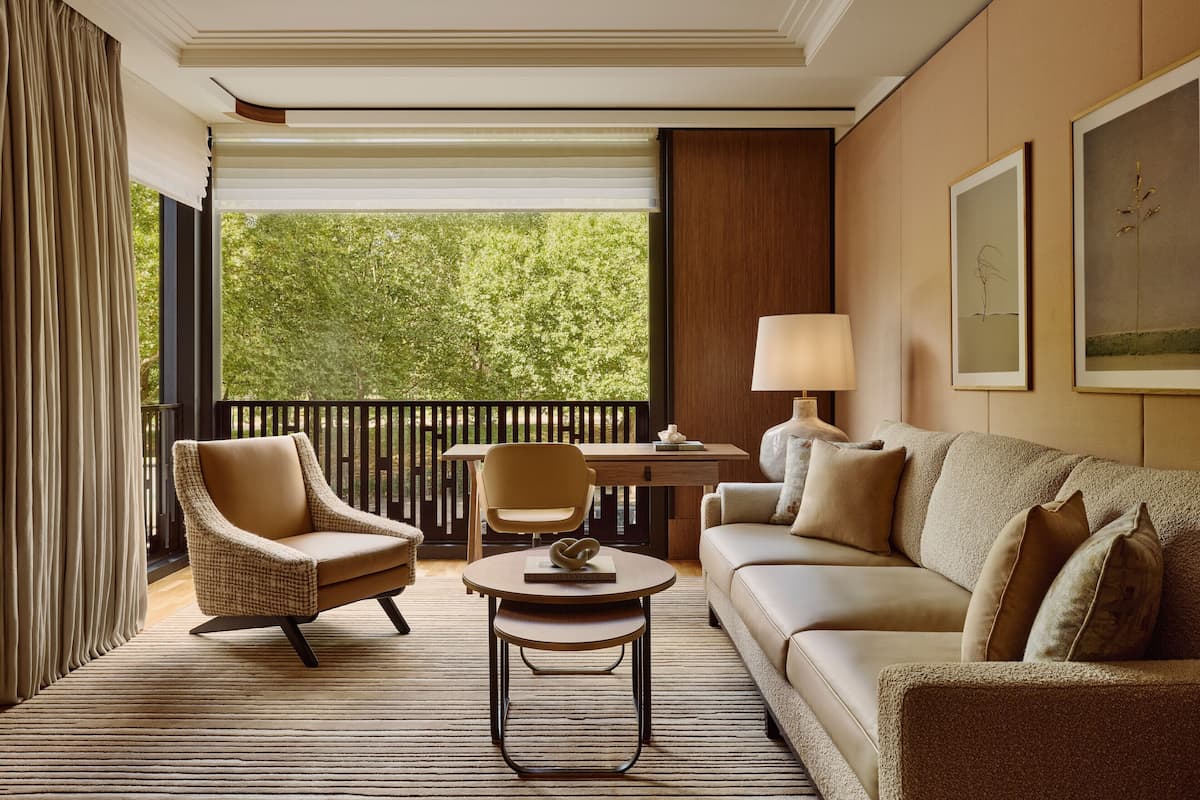 A room with a sofa, two armchairs, a small table, a lamp, and a view of greenery through a window.