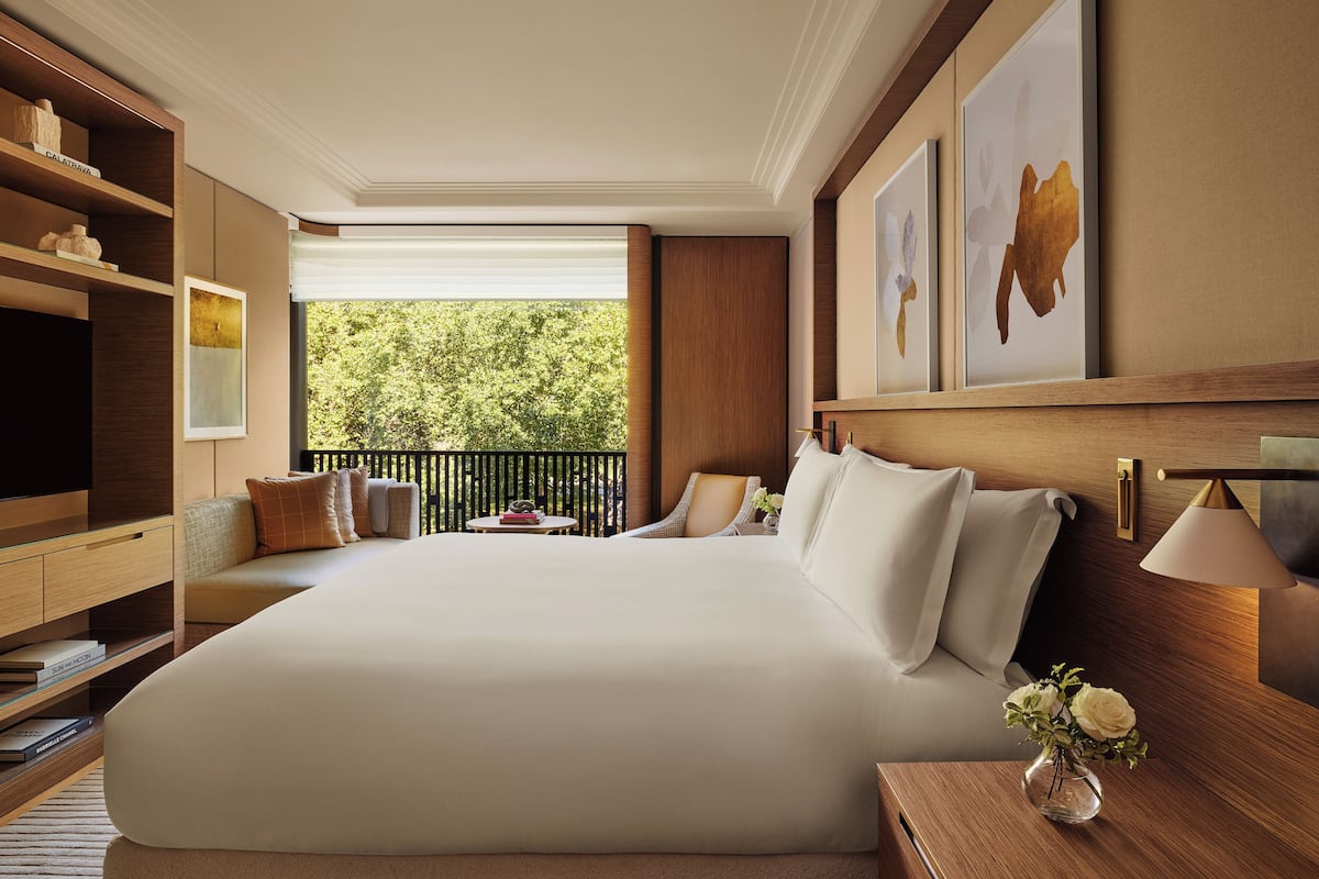 A spacious bedroom with a large bed, a wooden nightstand, a bedside table with a lamp, a bookshelf, and a view of greenery outside.