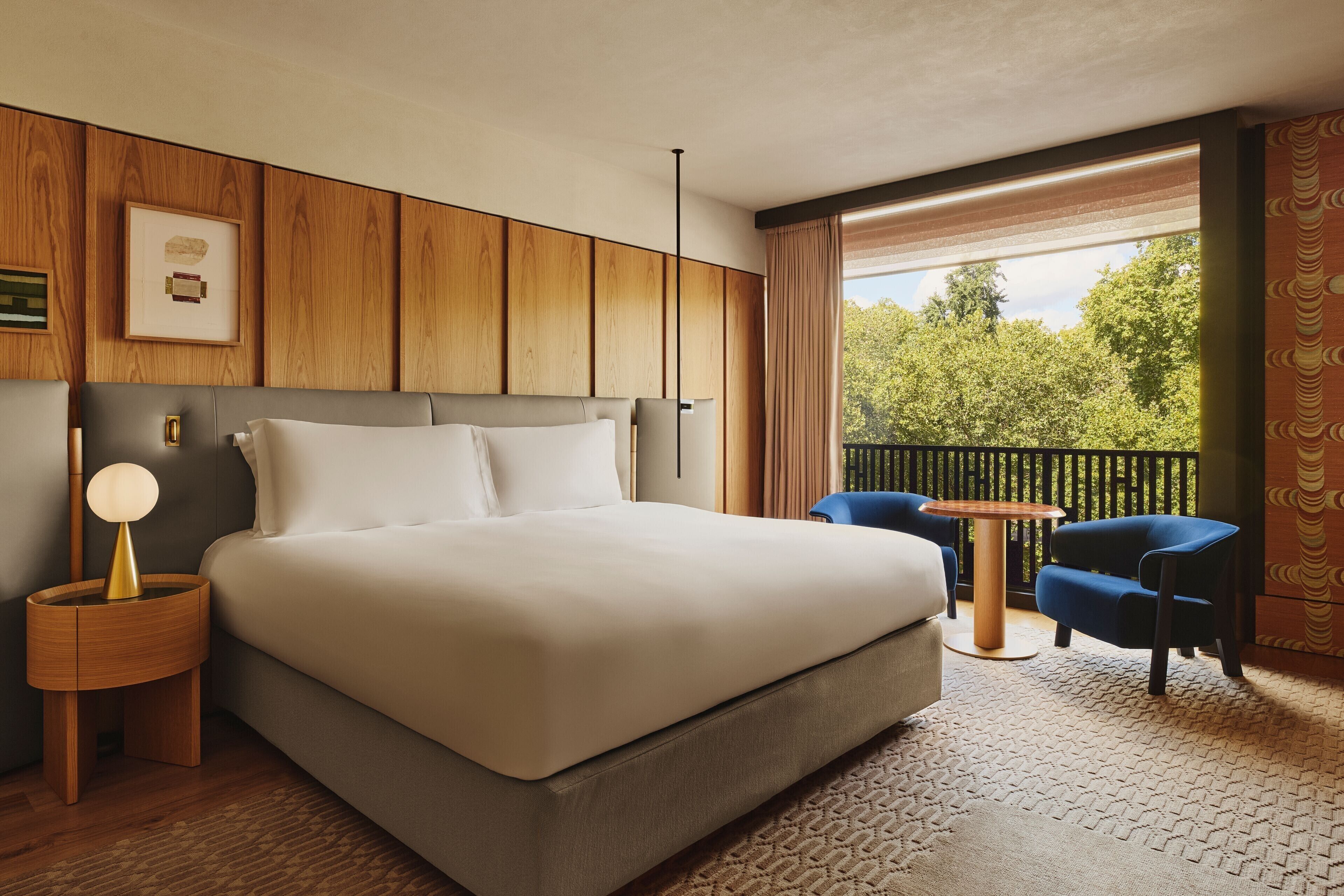 A hotel room with a large bed, a bedside table with a lamp, a small round table, two chairs, and a balcony with a view of trees.
